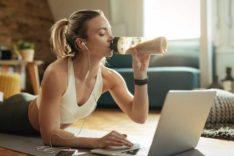 Eiwitten - Young athletic woman drinking protein shake while using laptop on the floor.
