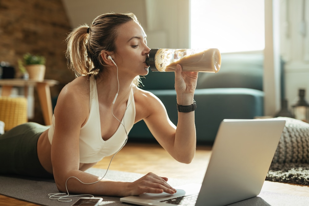 Eiwitten - Young athletic woman drinking protein shake while using laptop on the floor.