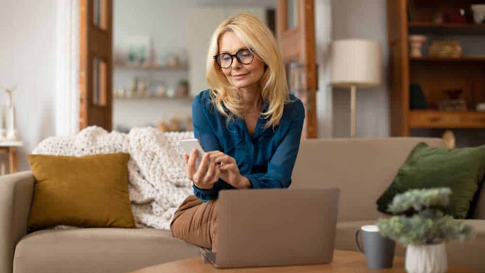 volwassen vrouw sms't op telefoon en werkt thuis op laptop