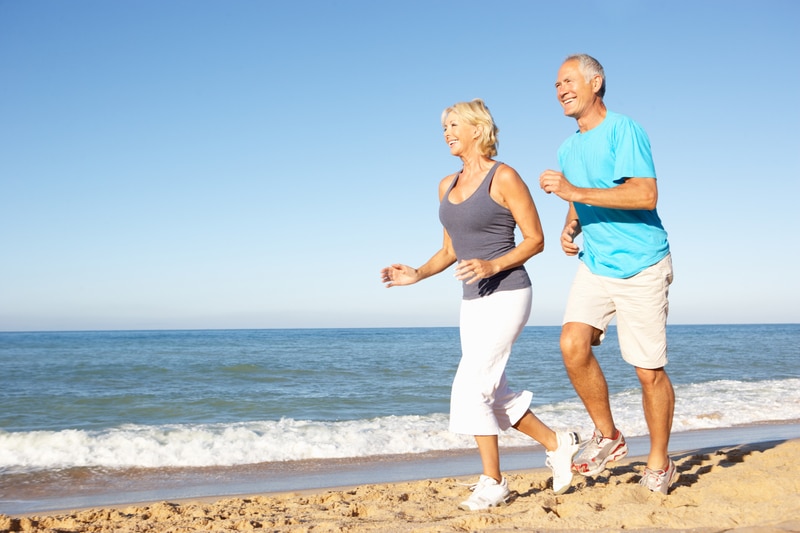 Oudere man en vrouw joggen samen langs de kustlijn op een zonnige dag, lachend en genietend van de frisse zeelucht.